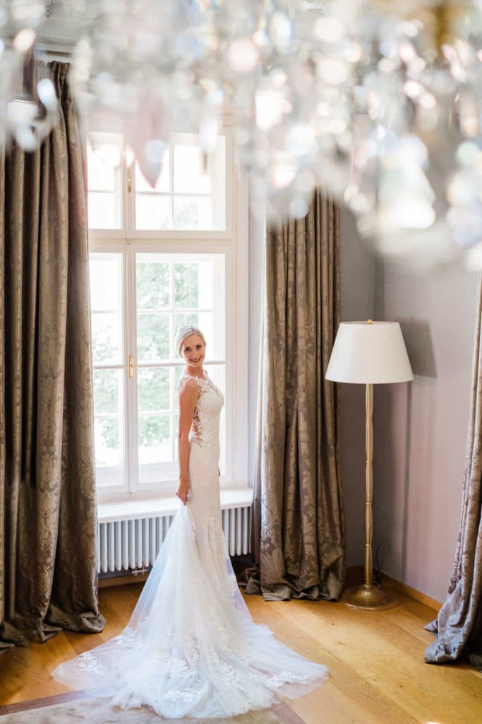 Braut steht im Brautkleid vor einem Fenster mit glamourösem Kristallluster